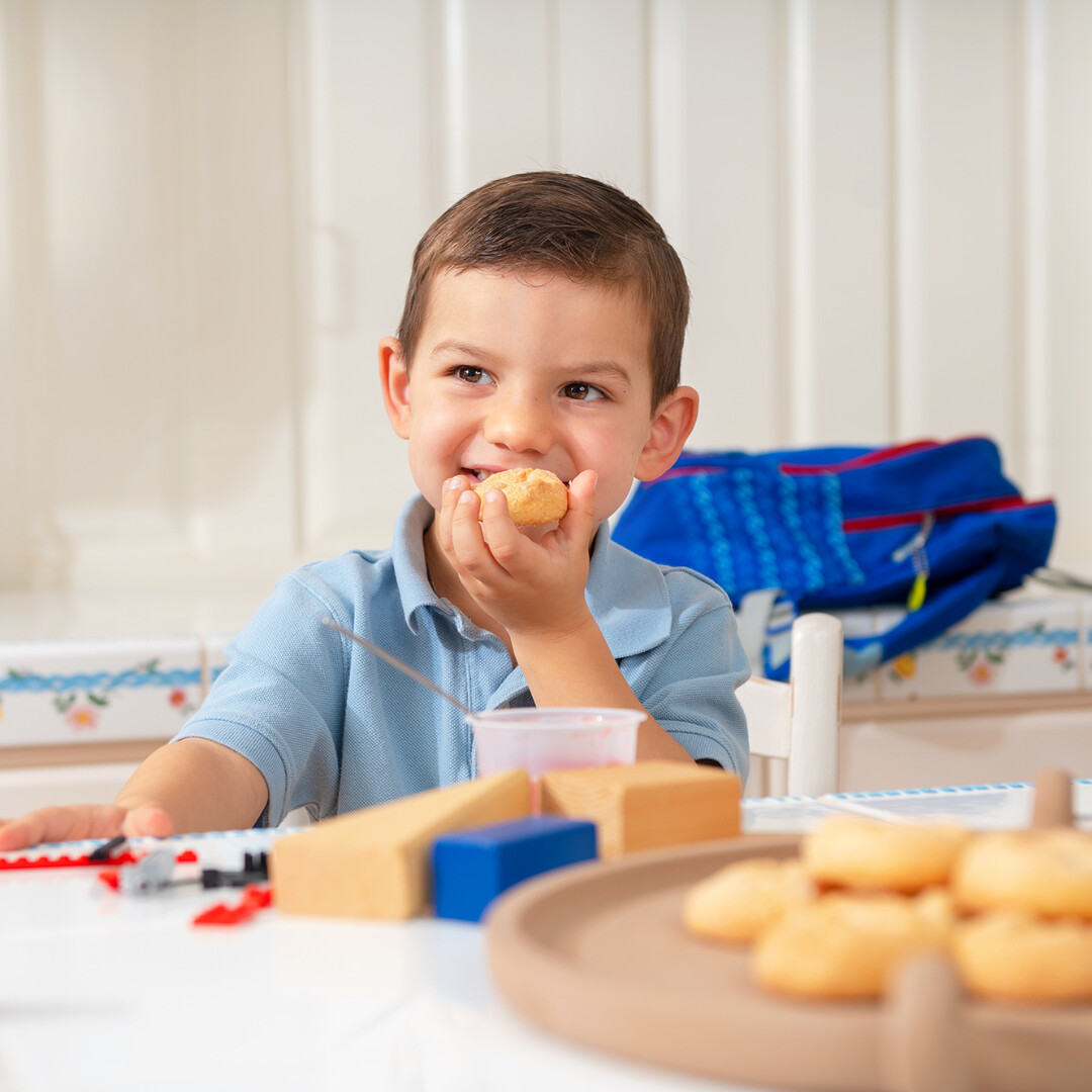 Joyful boy savoring a snack at a playful kitchen table with toys and pastries.