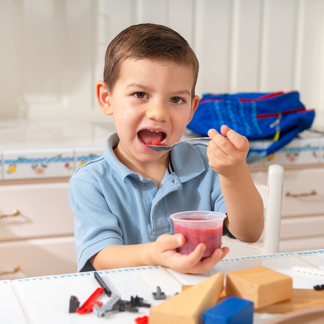 Cheerful boy enjoying gelatin dessert at kitchen table, surrounded by toys and snacks.