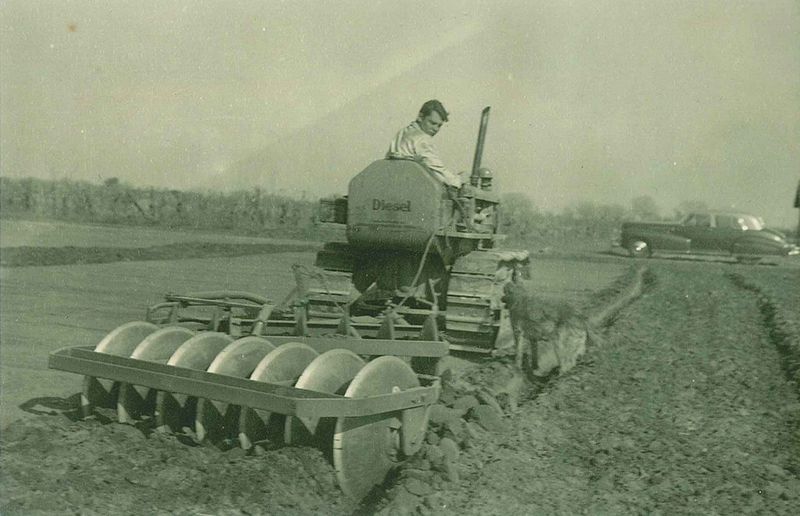 Farmer using vintage diesel tractor to plow soil, with dog and classic car nearby.